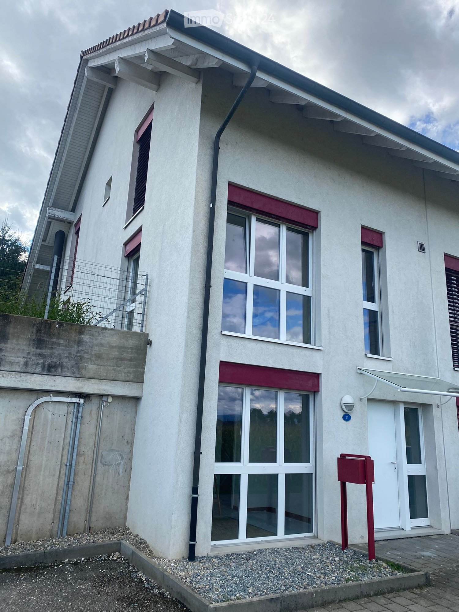 Two-story house with white walls, large windows, red shutters, and a small garden area with a metal fence.