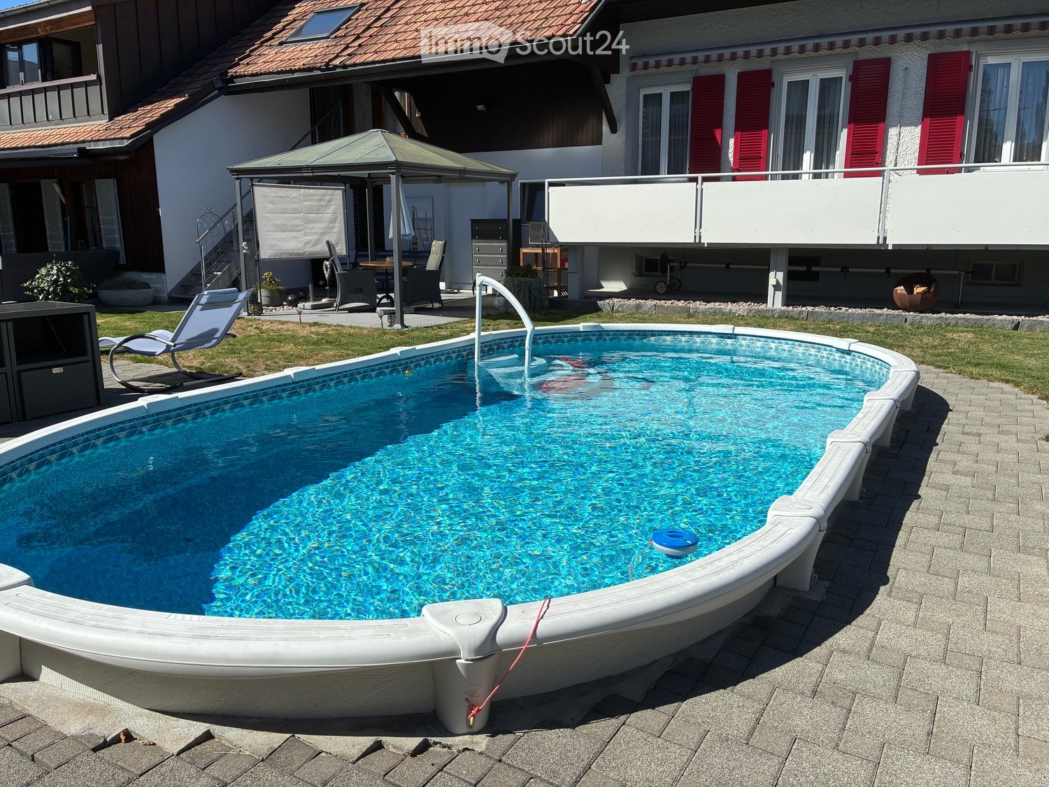 A private swimming pool with a modern design, surrounded by a paved patio and a well-maintained lawn. Nearby, there is a gazebo with outdoor furniture and a lounge chair.