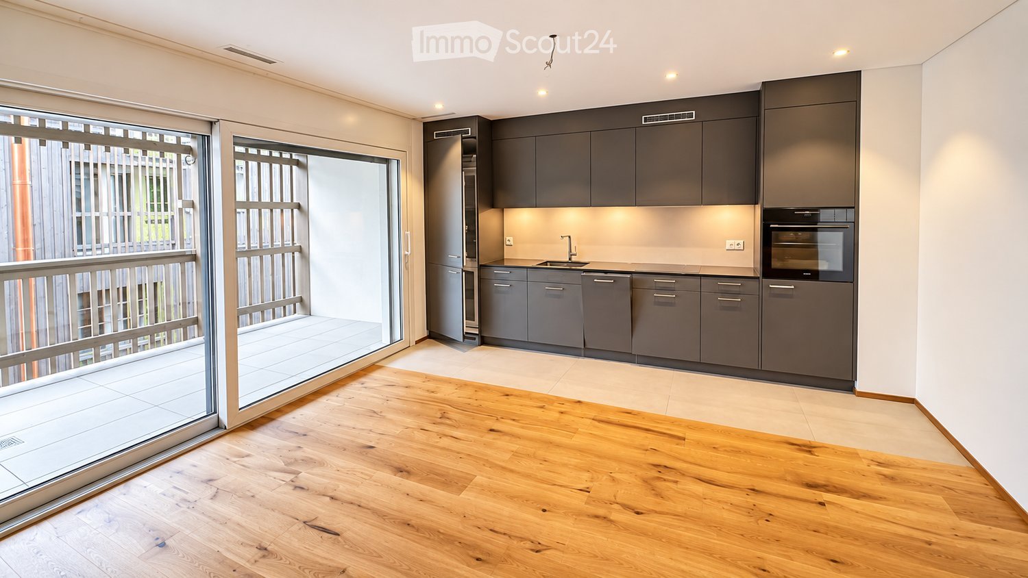 Modern kitchen with grey cabinets, a sink, and an oven, connected to a balcony with wooden flooring.