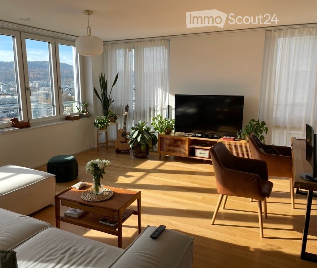 Bright living room with large windows offering a city view, featuring a grey sectional sofa, wooden coffee table, armchairs, a guitar, and several potted plants.