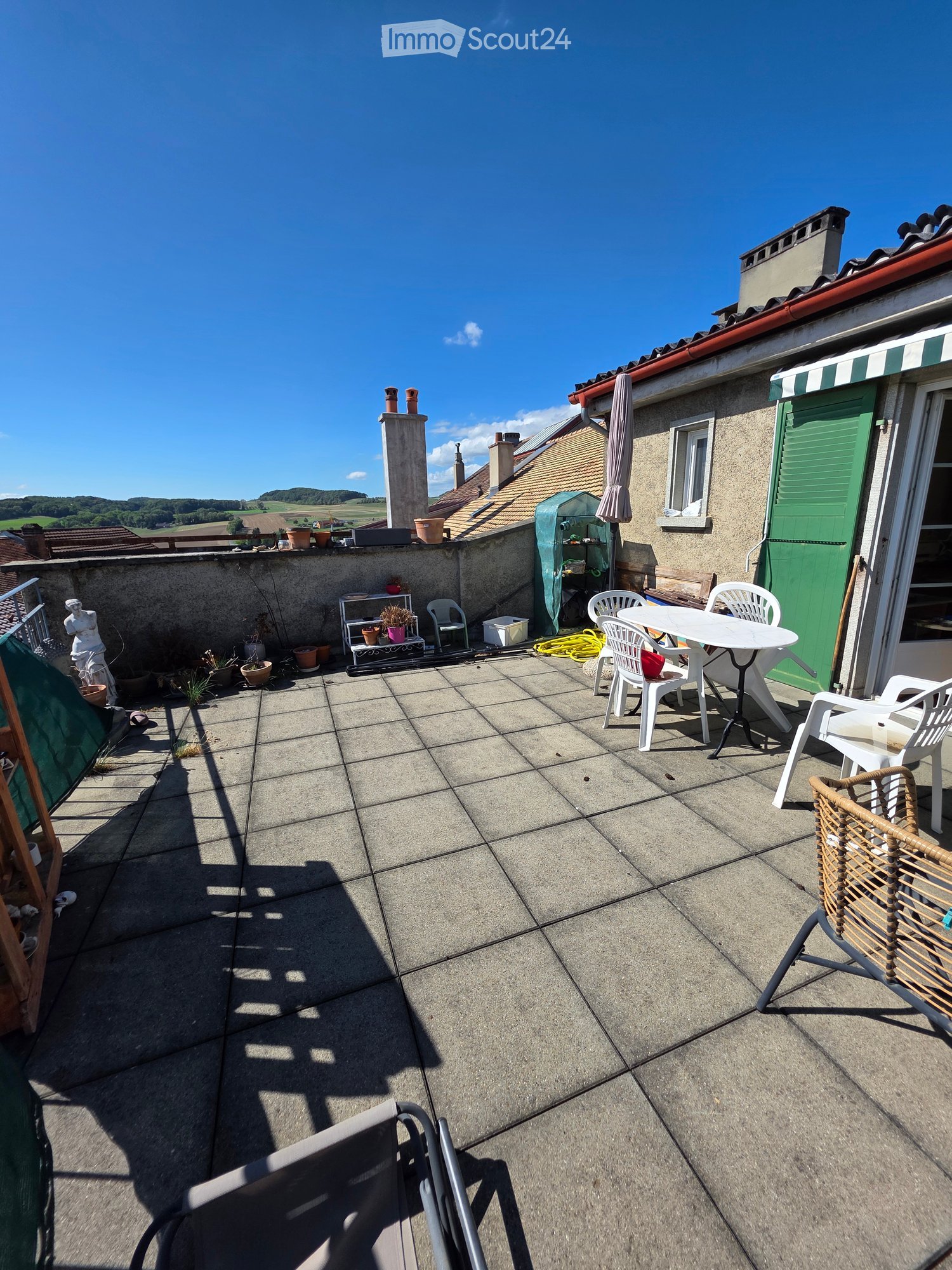 Roof terrace with concrete tiles, white plastic chairs and table, potted plants, and a green door to the building.