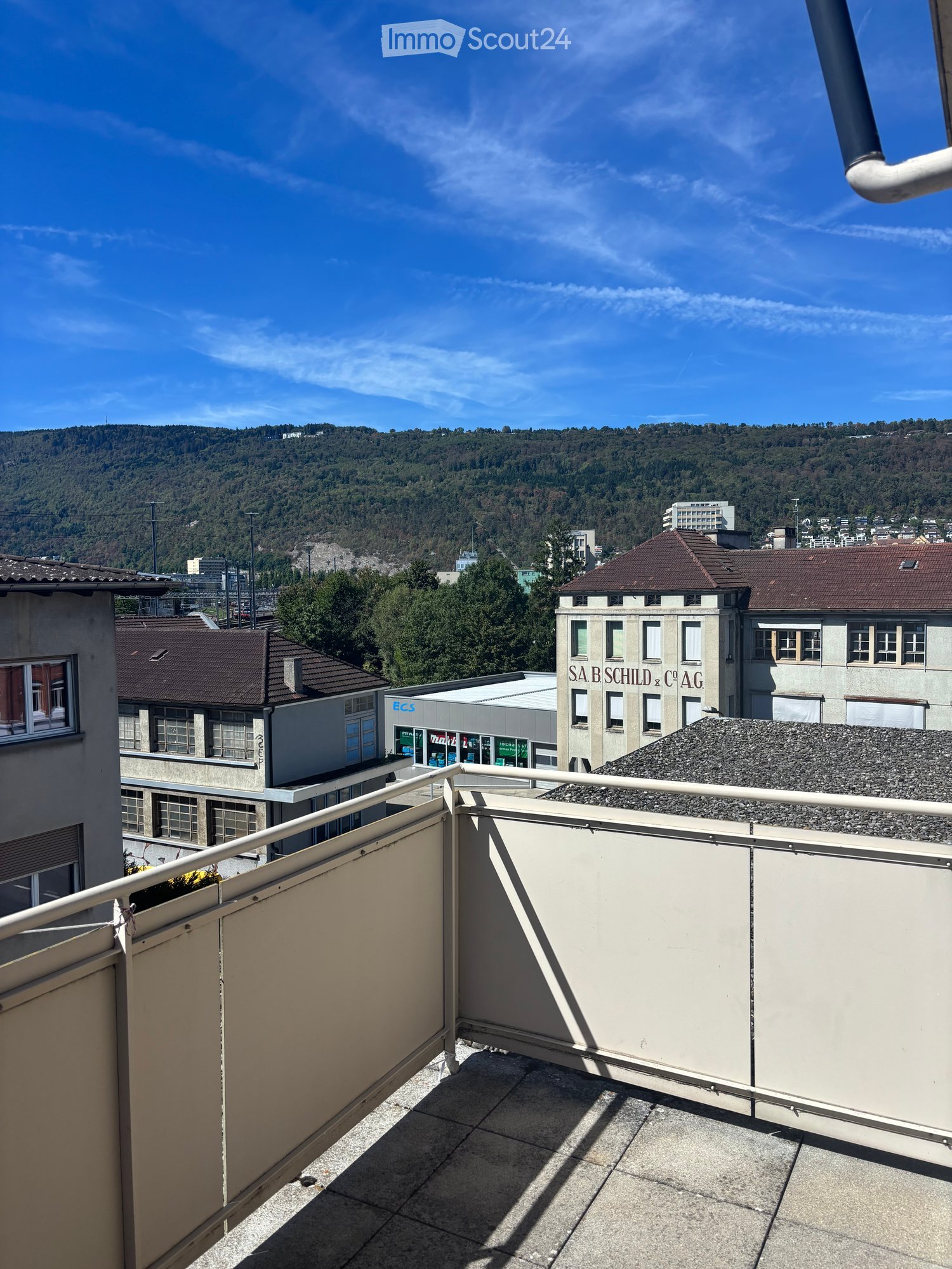 Balcony with a view of a cityscape and surrounding hills, featuring a metal railing and a gravel-covered roof area.