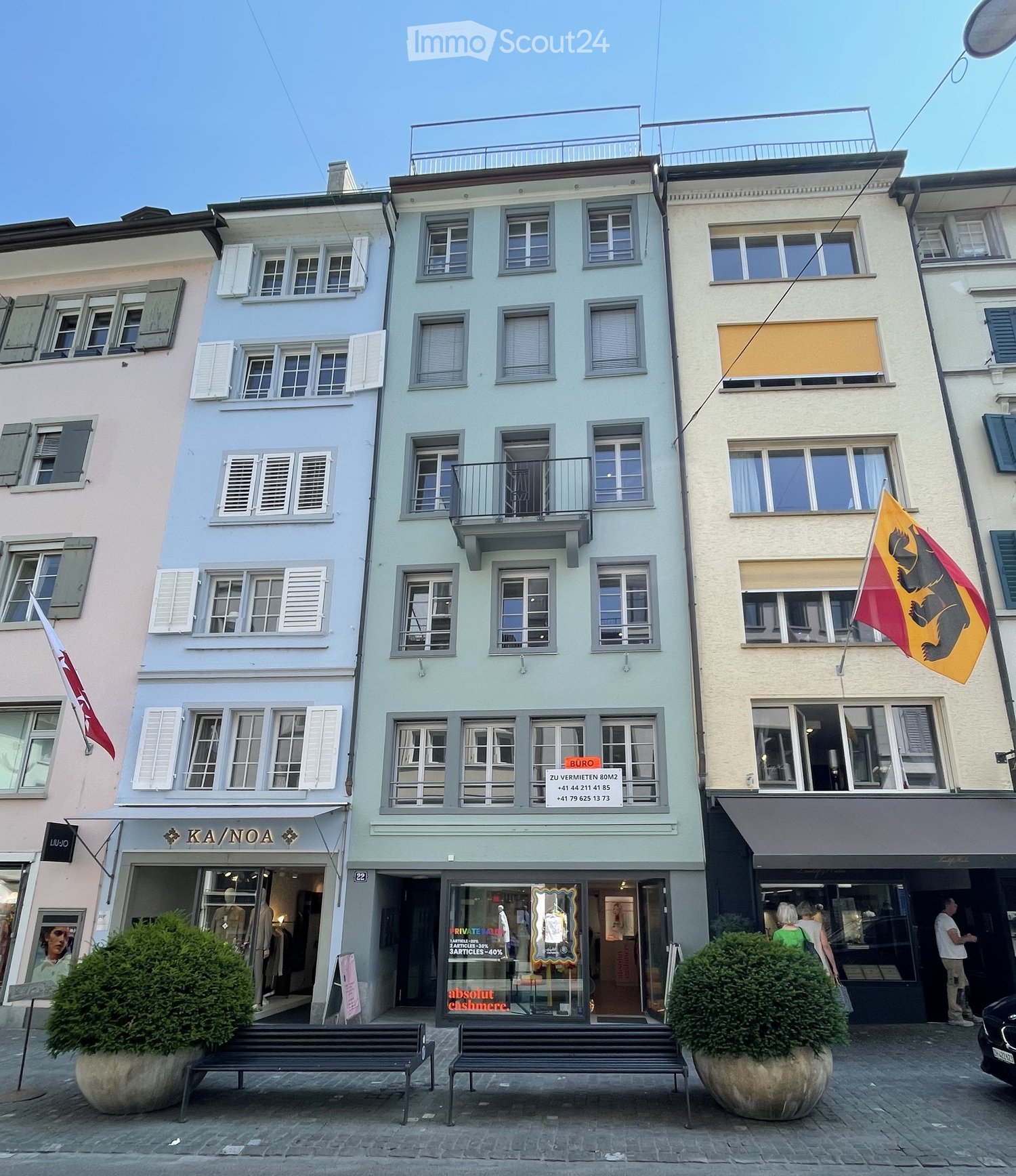 Three-story building with colorful facades, multiple windows, and a balcony on the second floor. The ground floor features storefronts with signage and mannequins in display windows.