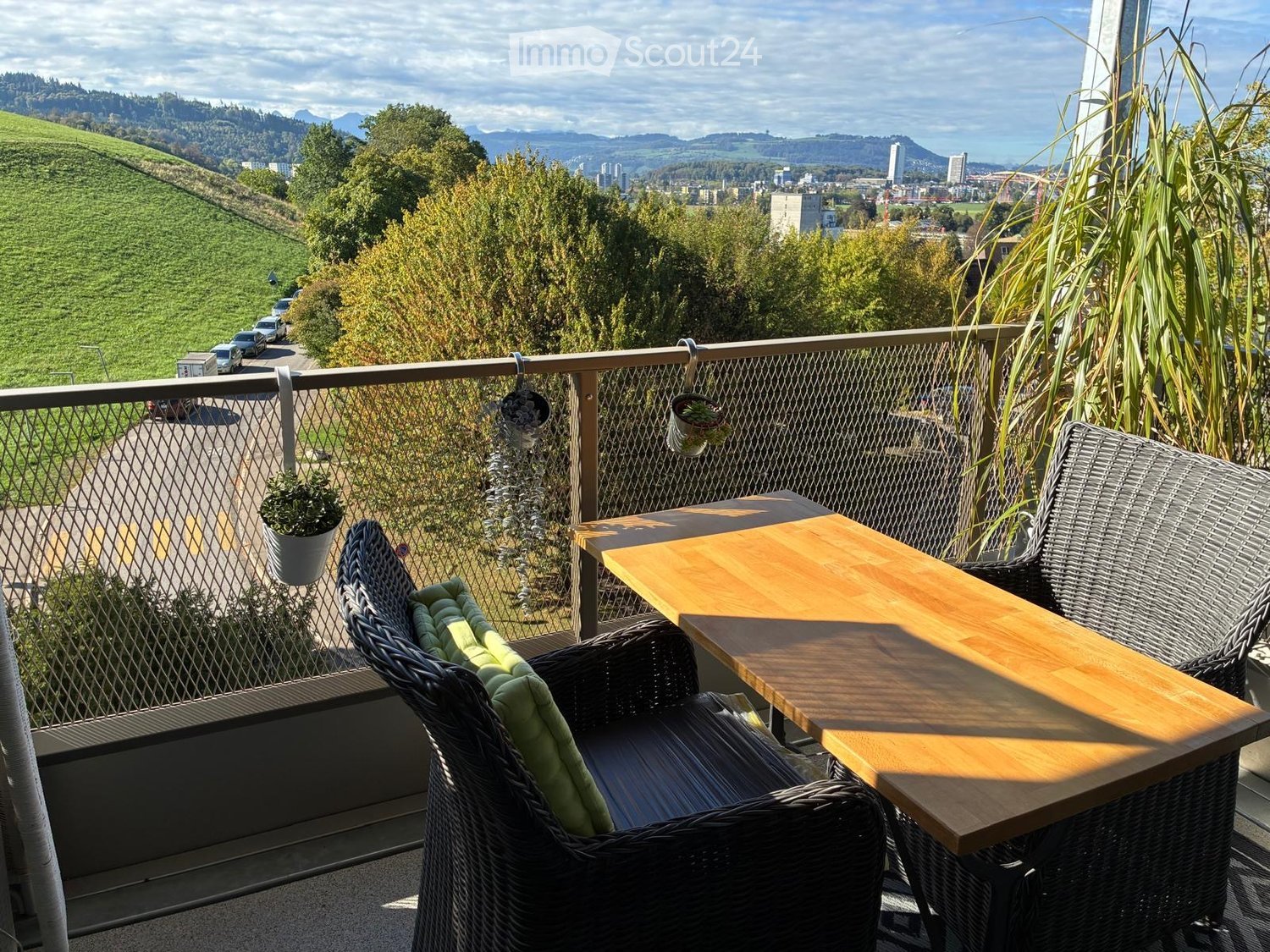 Balcony with a wooden table, wicker chairs, and potted plants, overlooking a scenic landscape with hills and a cityscape.