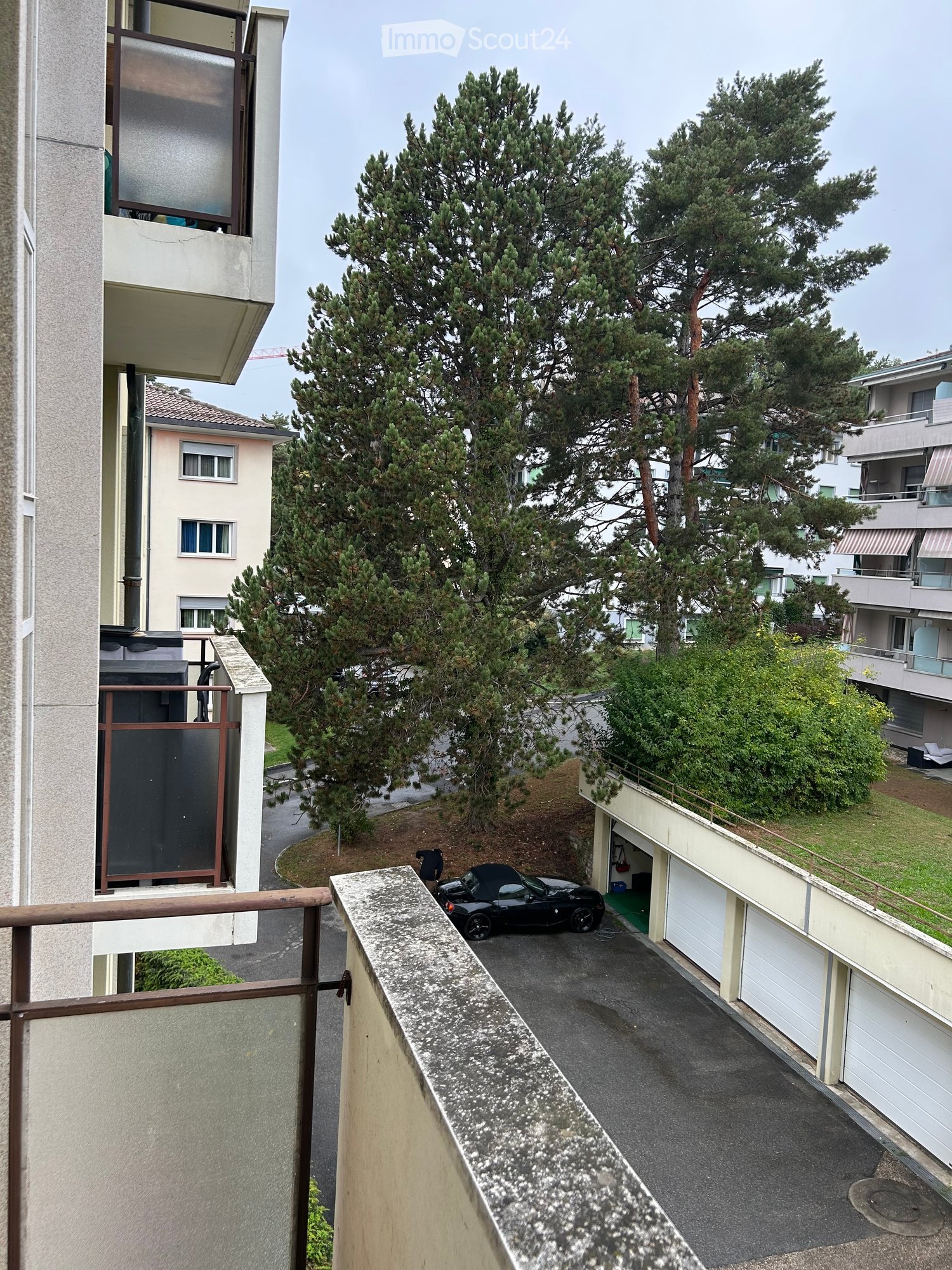 Balcony overlooking a parking area with a black car, surrounded by trees and neighboring buildings.