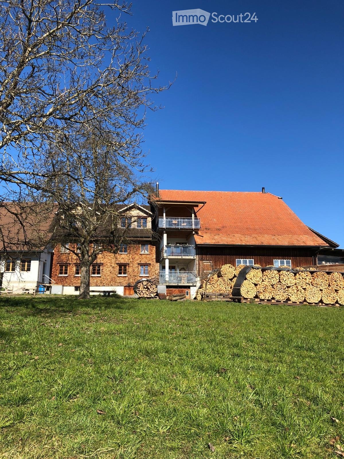 Two-story house with red-tiled roof, brick and wooden exterior, balcony, and stacks of firewood in front.