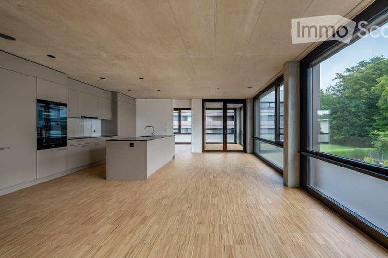 Open-plan kitchen and living area with light wood flooring, white cabinetry, and large windows offering a view of greenery. The space is unfurnished, with only built-in cabinetry and a kitchen island visible.