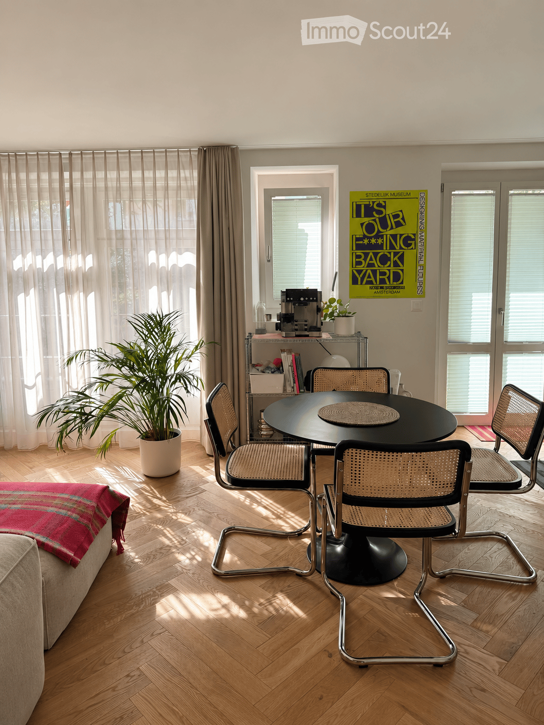 Bright living room with wooden herringbone flooring, large windows with sheer curtains, a round black dining table with four chairs, a sofa with a red and blue blanket, a potted plant, and a coffee machine on a shelf.