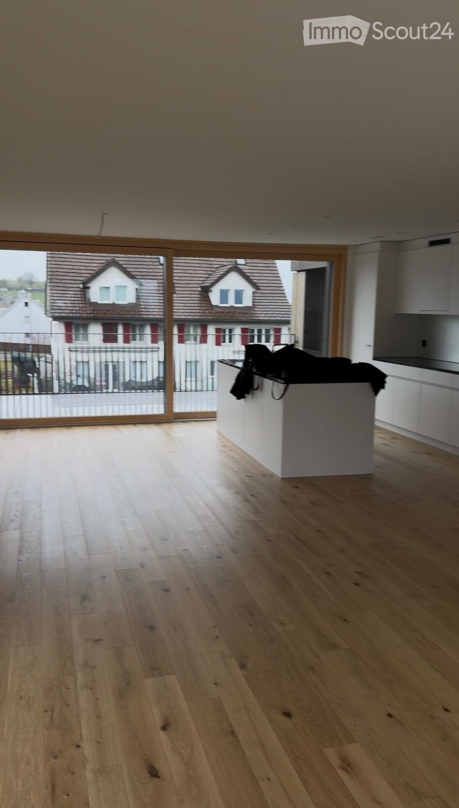 Empty room with light wooden flooring, white kitchen counter, and large windows overlooking a residential area.