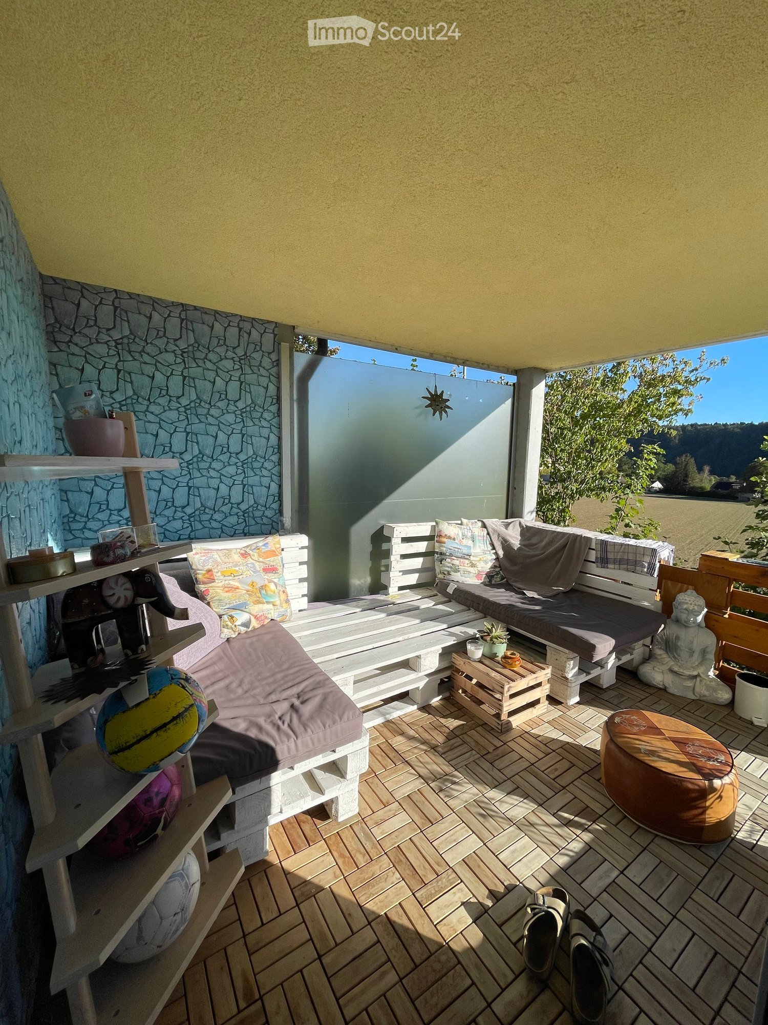 Balcony with wooden flooring, white wooden sofa, small table, round stool, shelf with decorative items, and a view of greenery.