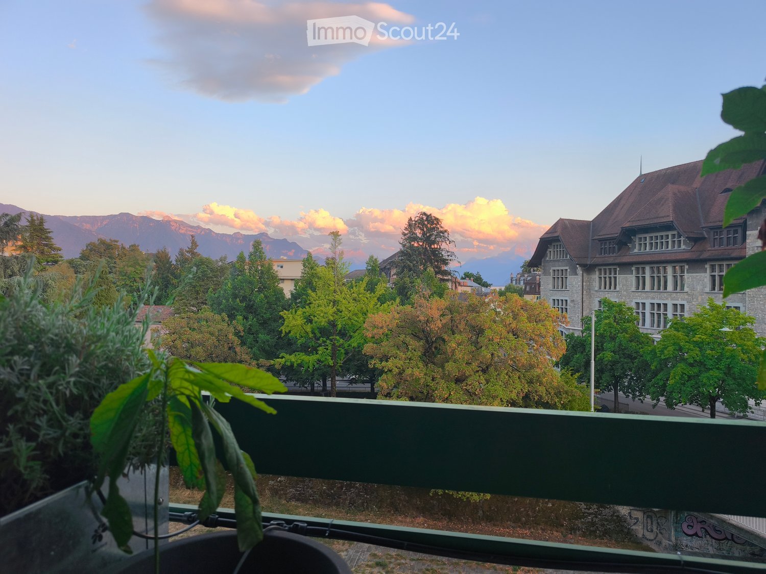 Balcony overlooking a tree-lined street with a large building and distant mountains under a colorful sunset sky.