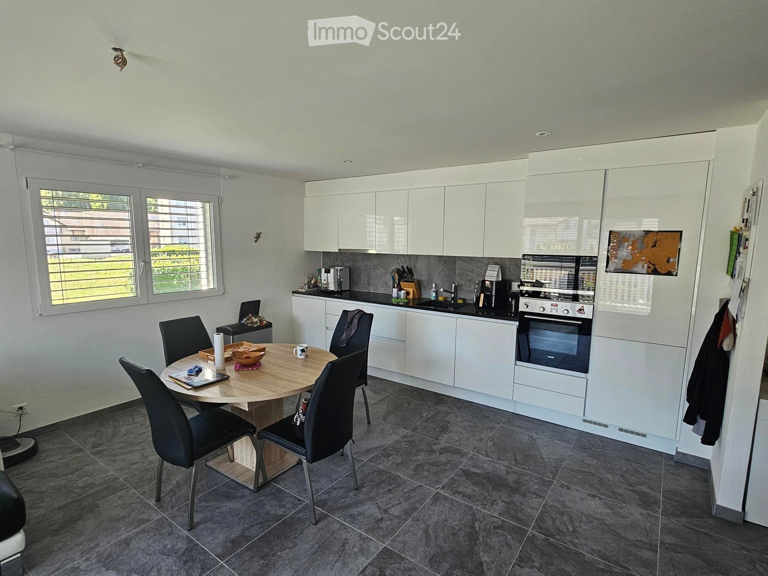 Modern kitchen with white cabinets, a round wooden dining table with four black chairs, and various appliances including an oven, coffee maker, and knife block. The space features large windows with blinds and a grey tiled floor.