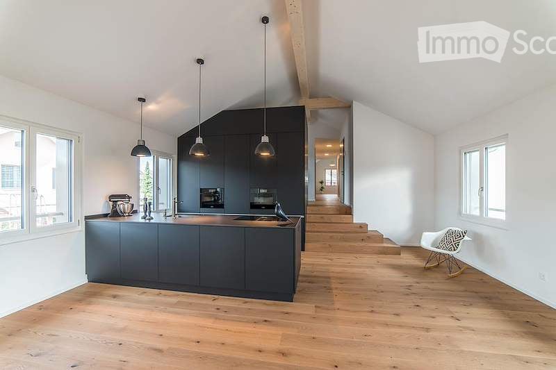 Modern kitchen with dark grey cabinetry, a central island, pendant lights, and a white chair near the staircase leading to another level.