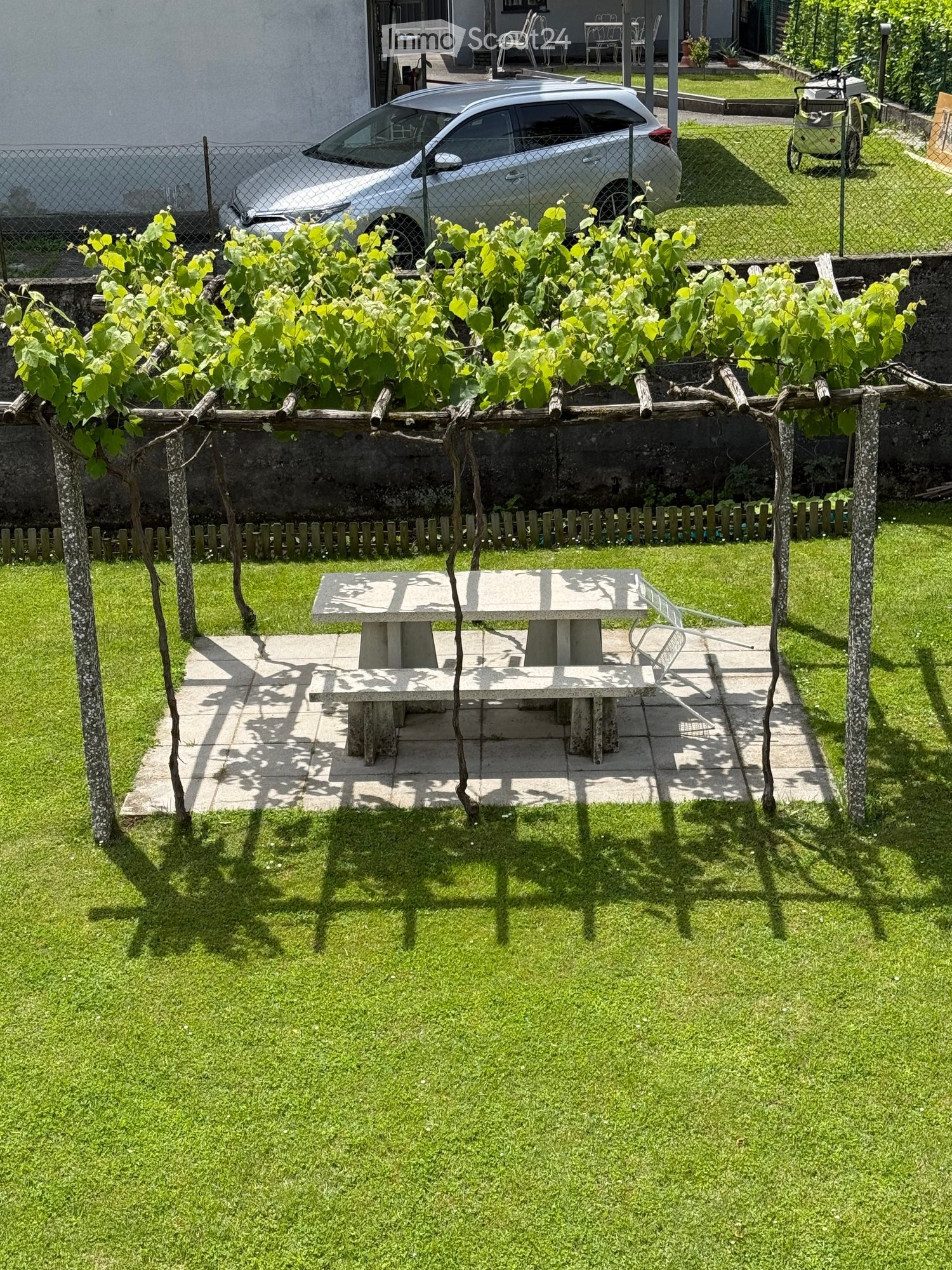 Well-maintained garden with a stone table and chair, surrounded by green grass and a wooden fence, with a grapevine trellis and a parked car in the background.