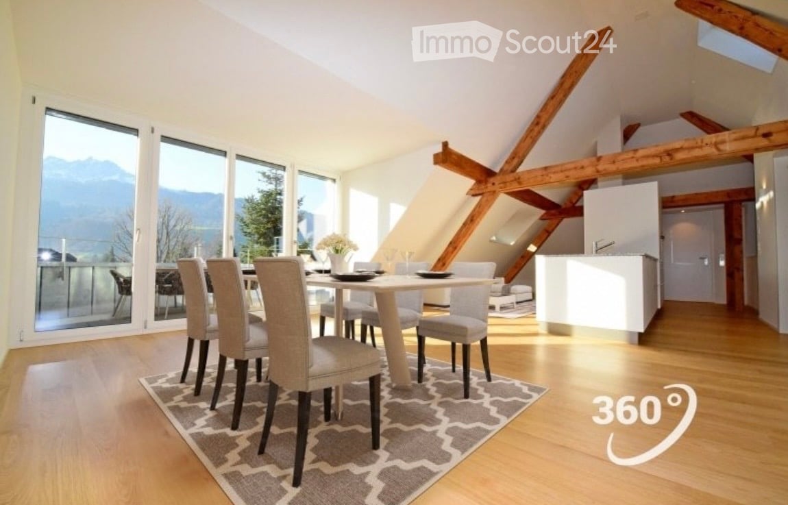 Bright dining area with a modern dining table and chairs, a vase with flowers, and a view of mountains through large windows. The room features wooden beams on the ceiling and a kitchen area visible in the background.