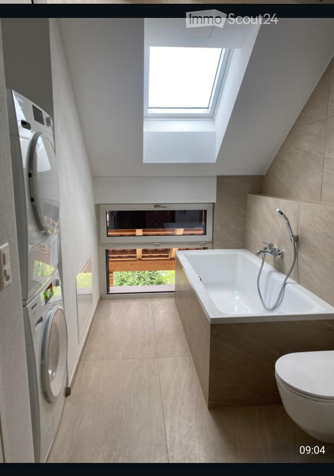 Modern bathroom with a bathtub, toilet, and washing machine. Features large windows and a skylight, with light-colored tiles and wooden accents.