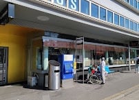 A modern building entrance with a yellow facade, glass windows, and a bus stop shelter nearby. There are a few people and a bicycle visible.