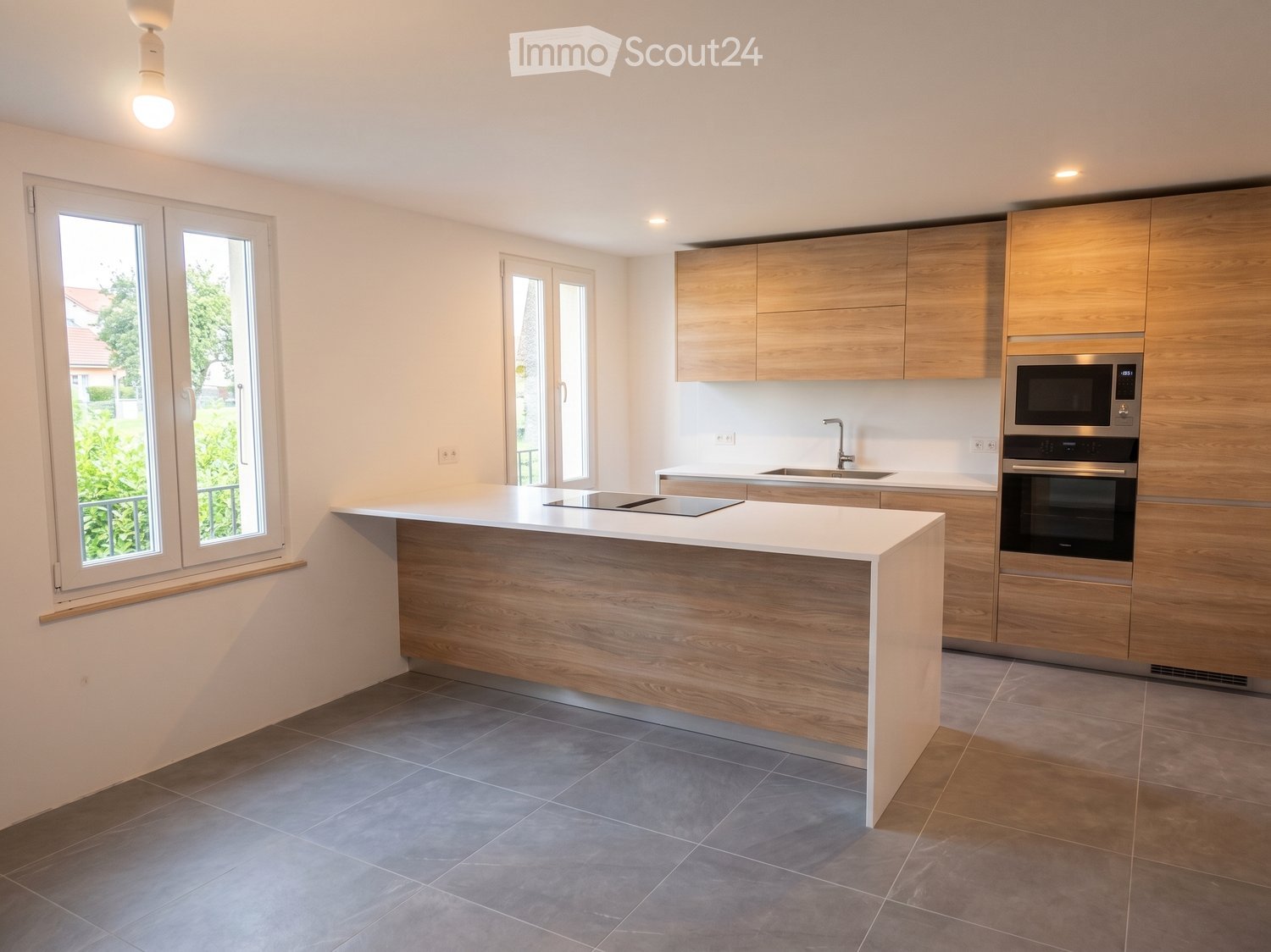Modern kitchen with light wooden cabinets, white countertops, and stainless steel appliances including a microwave and oven. Features a central island with a built-in induction cooktop and ample natural light from large windows.