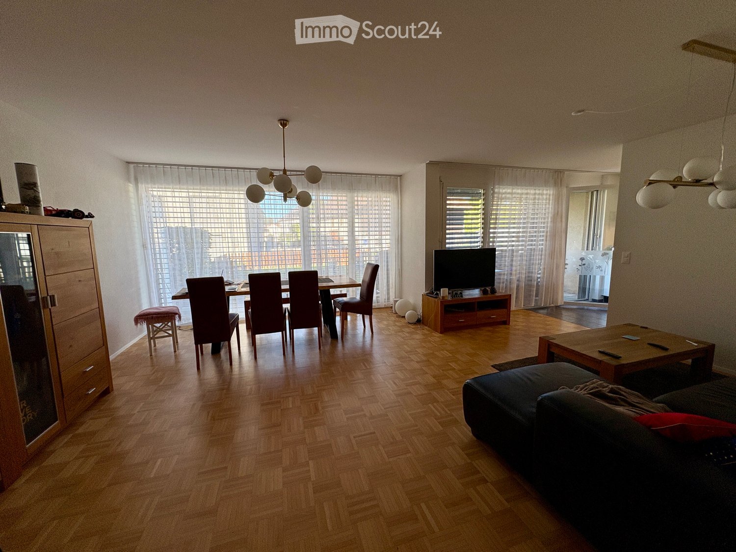 Open-plan living and dining area with wooden flooring, large windows, a sofa, coffee table, dining table with chairs, TV stand, and a cabinet.