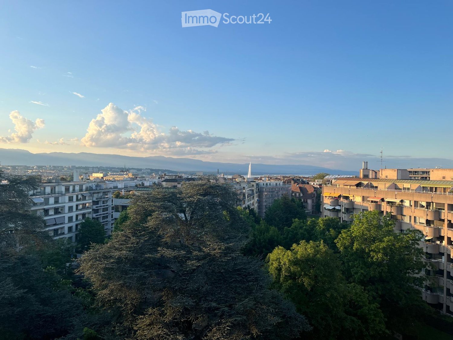 Panoramic view of a cityscape with multiple residential and commercial buildings, surrounded by trees and a clear blue sky with distant mountains.