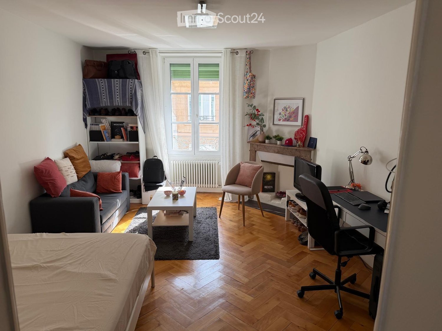 A multi-functional room with a bed, sofa, coffee table, desk, and chair, featuring wooden flooring and large windows with white curtains.