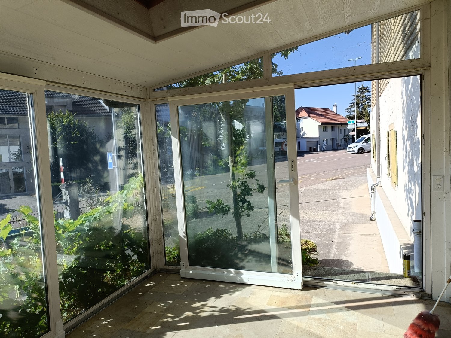 Bright, enclosed balcony area with large glass sliding doors, tiled floor, and visible outdoor greenery and street view.