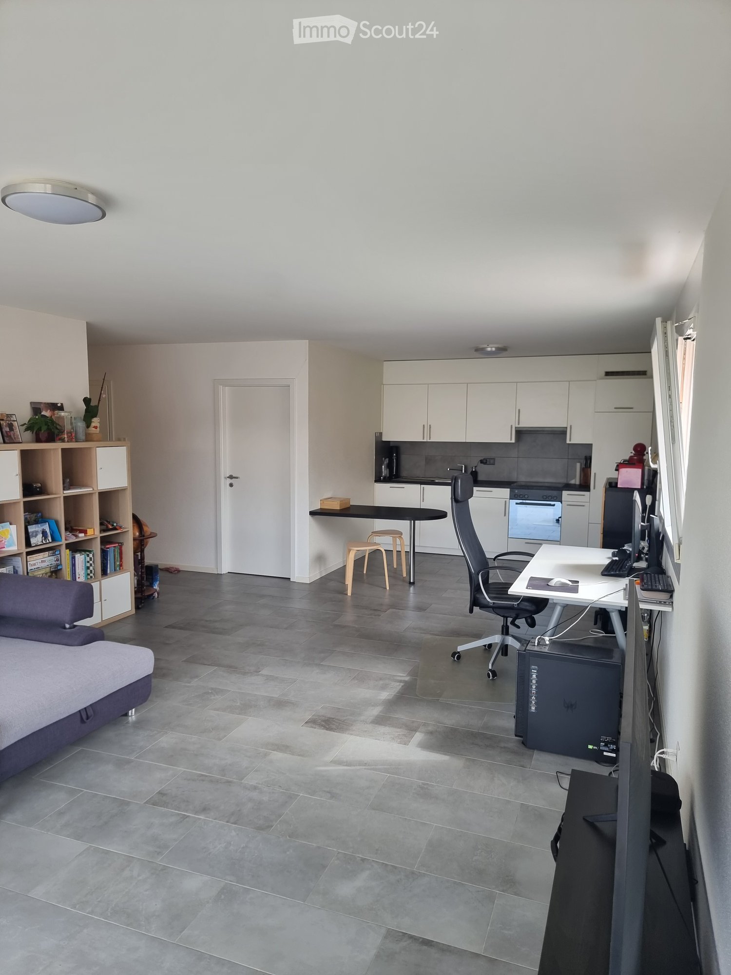 Open-plan living and working space with a grey sofa, office chair, desk, small table with stools, kitchen area with cabinets, oven, and fridge, bookshelf with books and decor, and a large screen with computer setup.