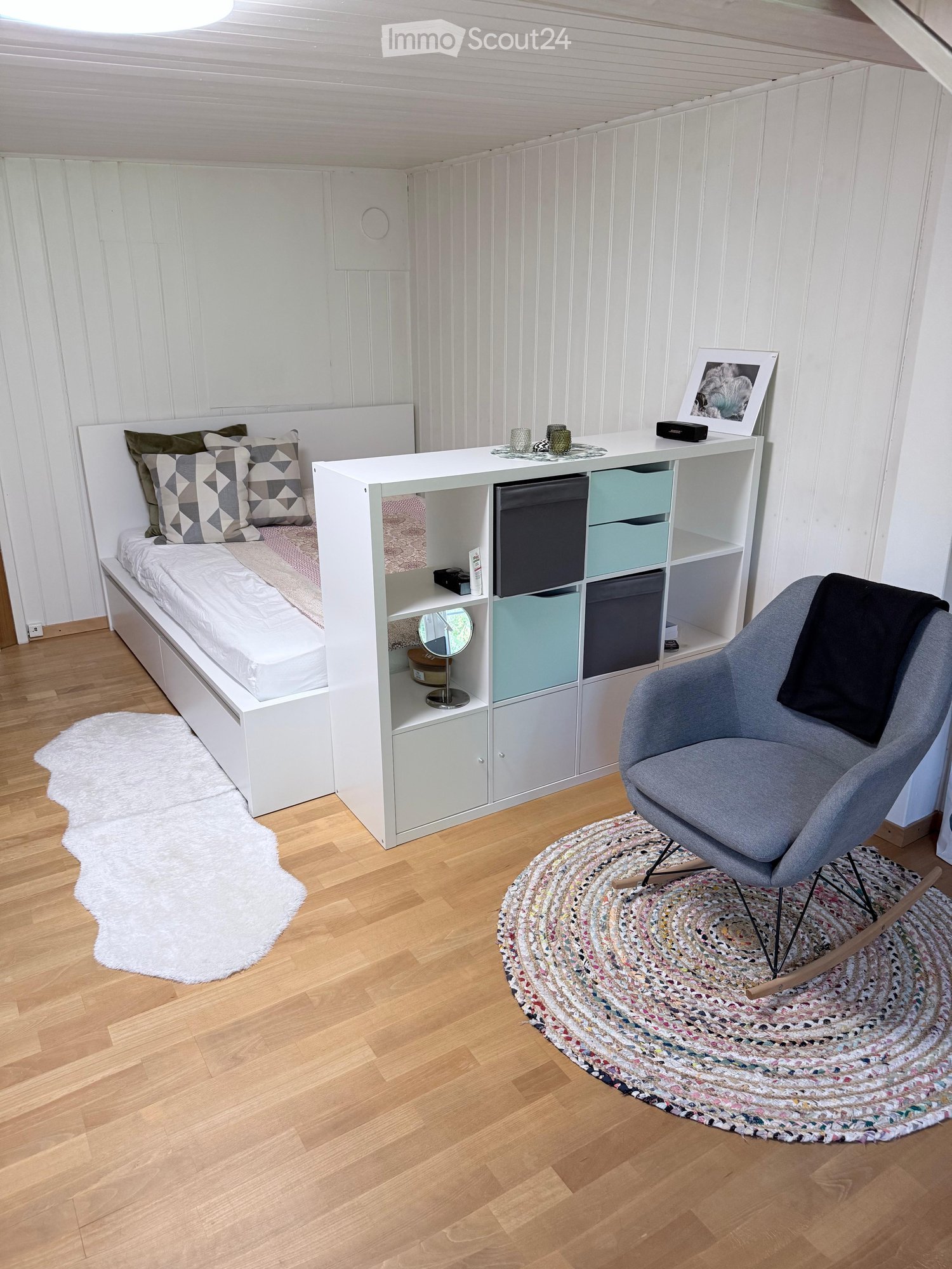 Cozy bedroom with a white bed, patterned pillows, a grey armchair, a white shelf unit with storage compartments, a round woven rug, and a white shaggy rug near the bed.