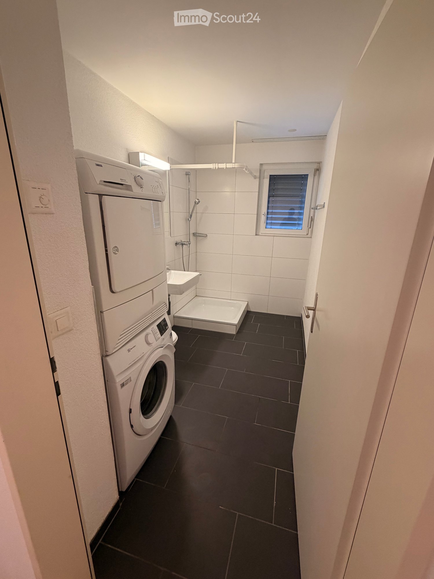 Modern bathroom with black tiled floor, white tiled walls, a stacked washing machine and dishwasher unit, a shower stall, and a small sink.