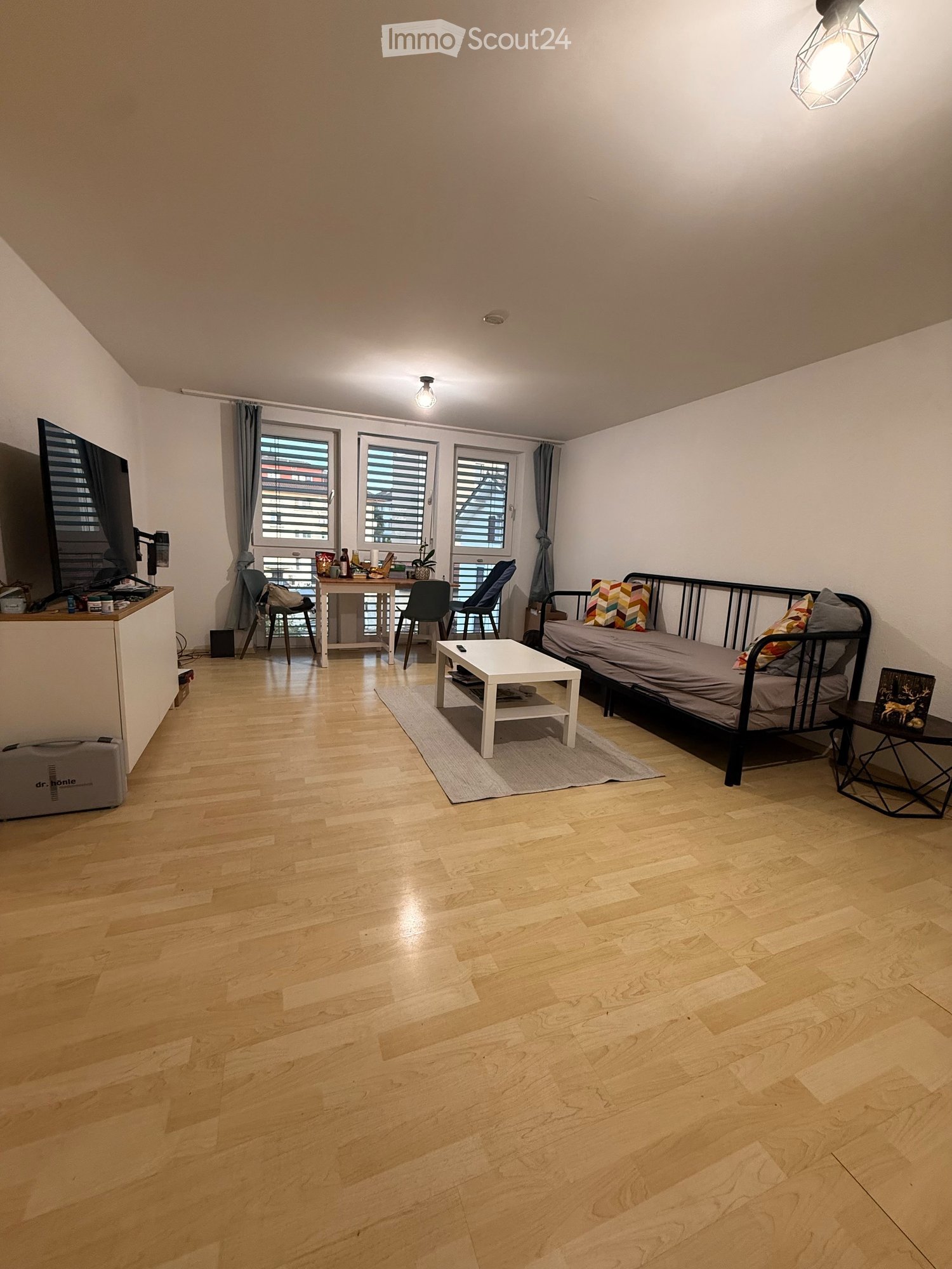 A modern, multi-functional room with a sofa bed, coffee table, small table, chairs, and a TV screen. The room features wooden flooring, large windows with blinds, and a cozy layout.
