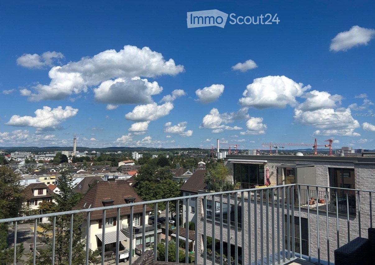 Balcony overlooking a cityscape with residential buildings, trees, and distant construction cranes under a partly cloudy sky.