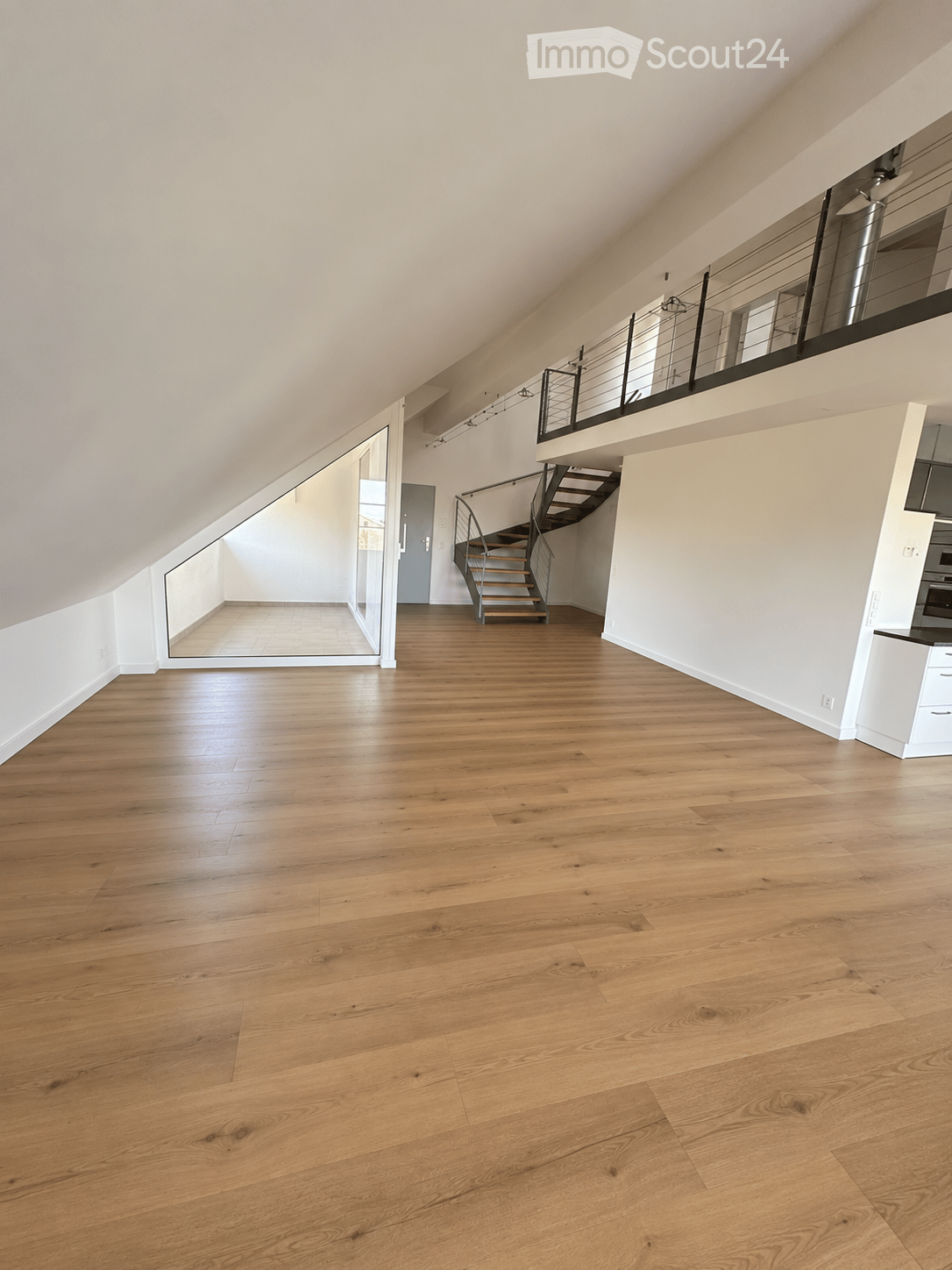 An empty, unfurnished room with light wooden flooring, white walls, and a staircase leading to an upper level. The space features a modern design with large windows and a glass door leading to another area.
