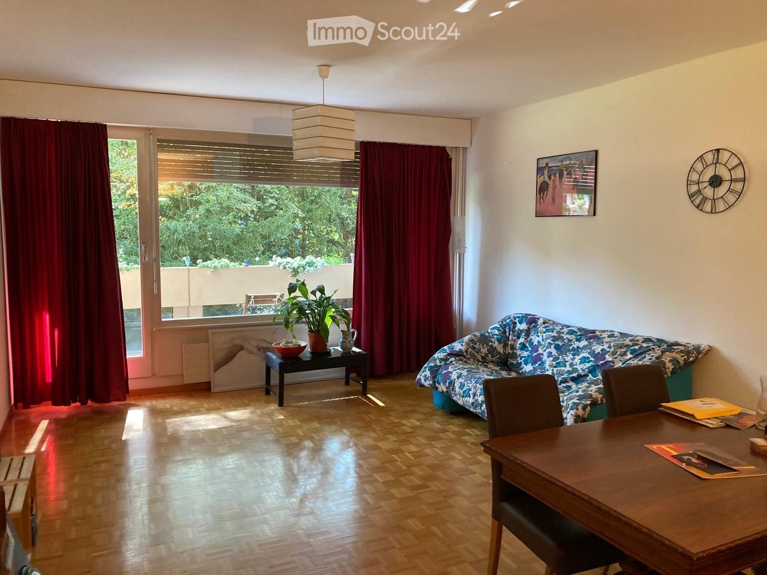 Living room with wooden flooring, large window with red curtains, sofa, small table with plant, dining table with chairs, and artwork on the wall.