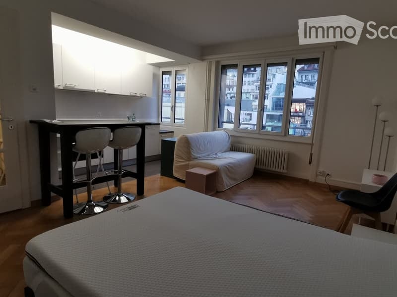A modern, open-concept living space with a bed, sofa, and kitchen area. The room features large windows with city views, wooden flooring, and contemporary furniture including a sofa, a small table, and bar stools.