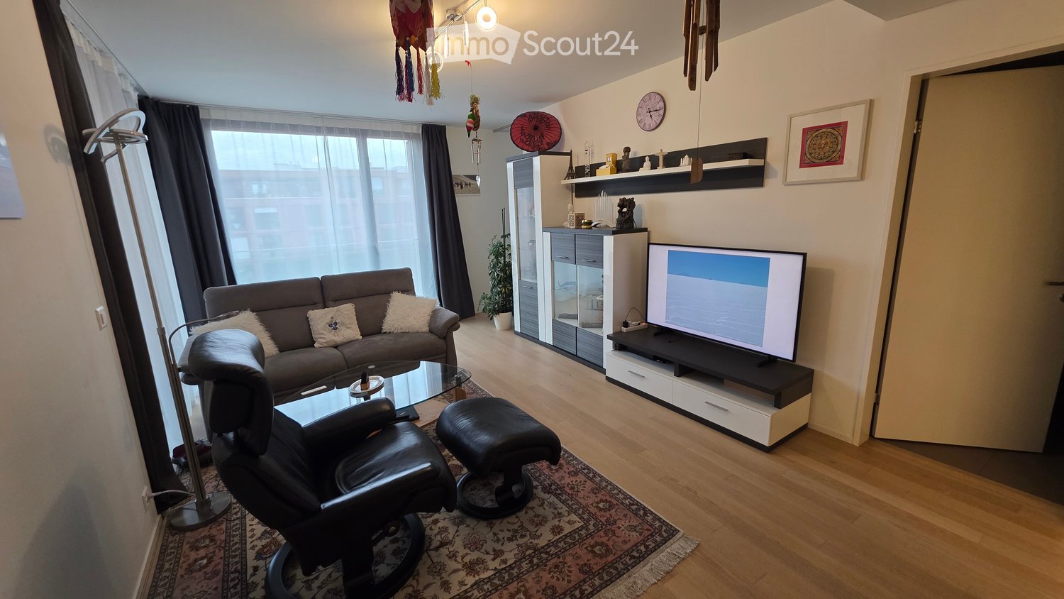 Modern living room with a grey sofa, glass coffee table, leather chair, ottoman, TV, cabinet, and decorative items.