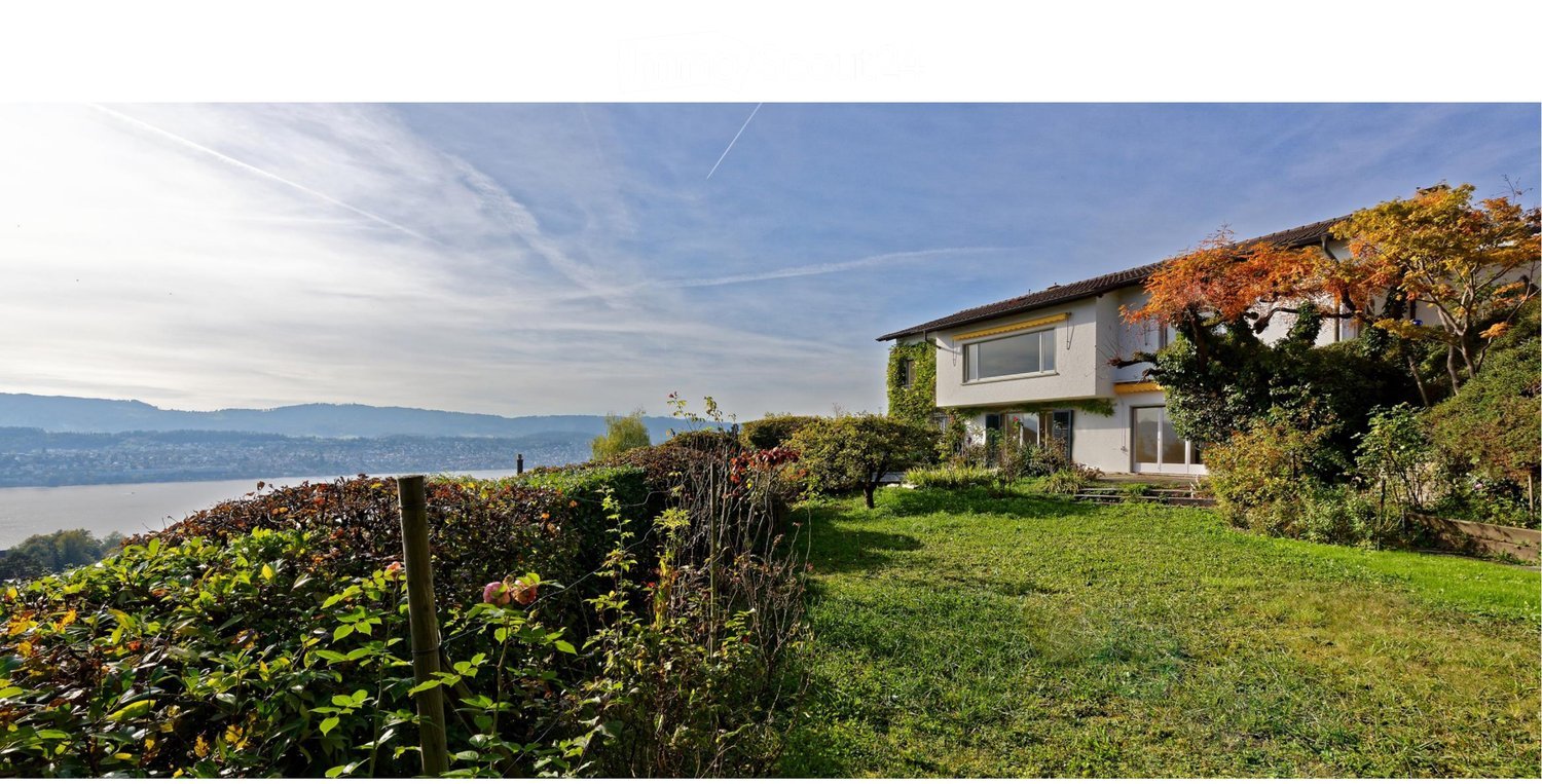 A two-story house with white walls and large windows, surrounded by a well-maintained garden and trees, overlooking a scenic lake and distant mountains.