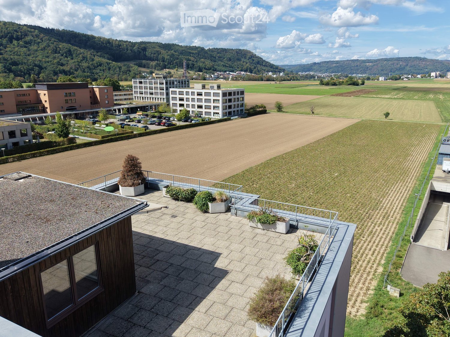 Aerial view of a modern building complex with a spacious rooftop terrace, surrounded by open fields and distant hills.