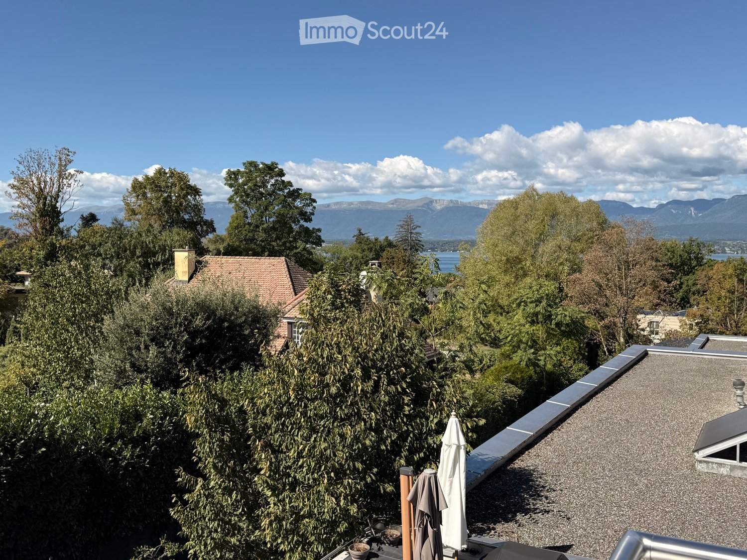 A rooftop terrace with a view of a lake and mountains, surrounded by trees and a house with a red-tiled roof.