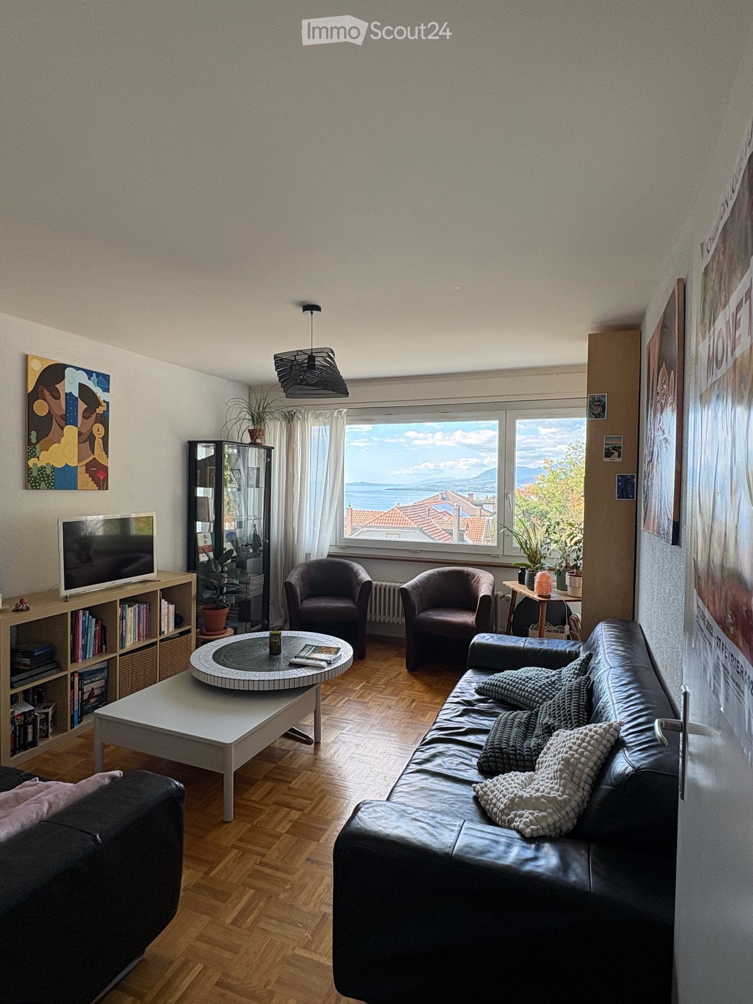 Modern living room with wooden flooring, large windows offering a scenic view, a black leather sofa with blankets, a white coffee table, two armchairs, a bookshelf, and a television.