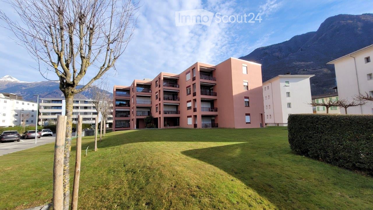 Modern multi-story residential building with pink facade and balconies, surrounded by green lawn and trees, with mountains in the background.