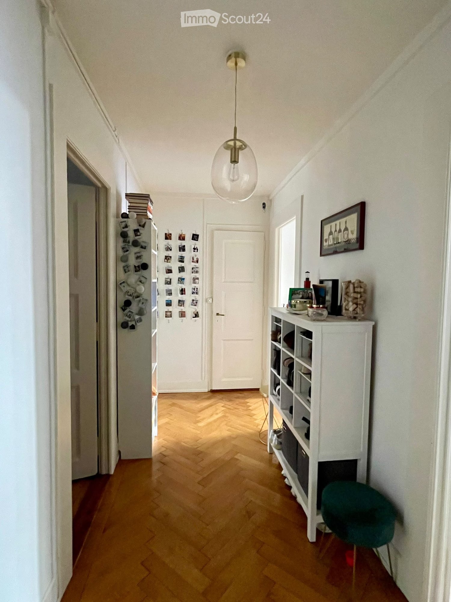 A narrow hallway with wooden herringbone flooring, white walls, and a modern pendant light fixture. A white shelving unit with books and decorative items is visible on the right, along with a green chair and a framed picture on the wall.
