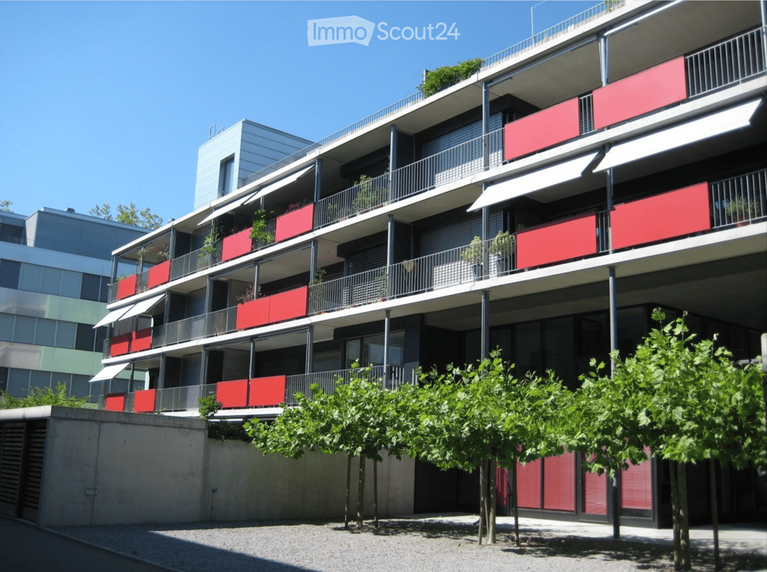 Modern multi-story residential building with red balconies, metal railings, and greenery in front.