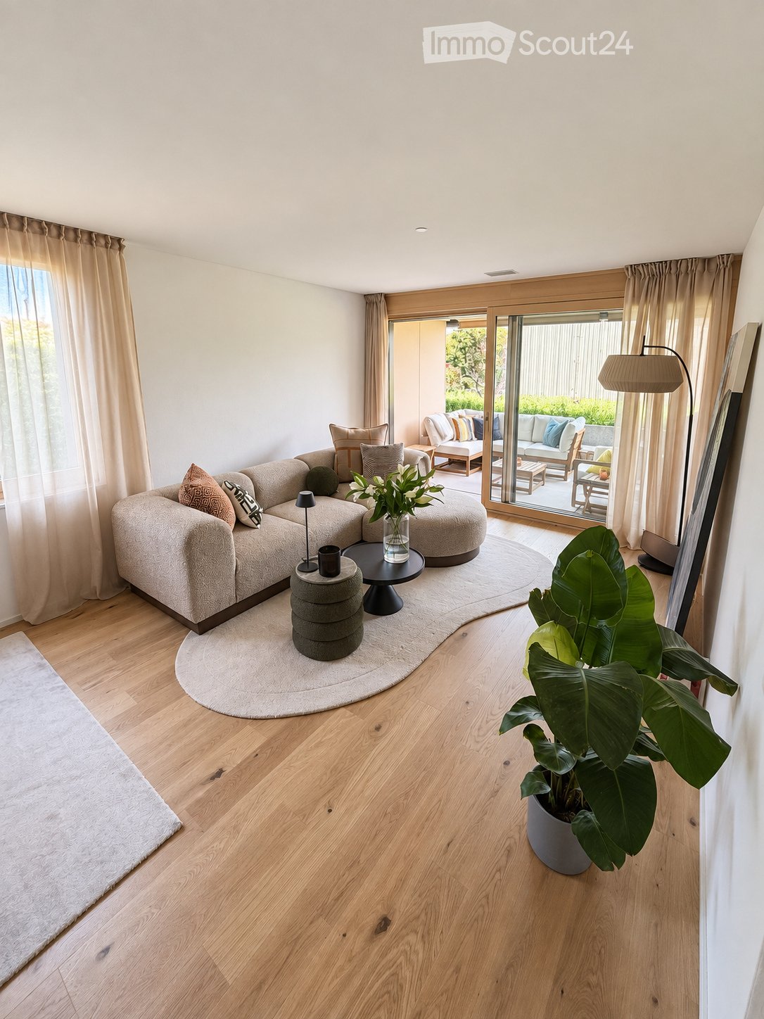 Modern living room with light wooden flooring, large sectional sofa, coffee table, floor lamp, and a large potted plant near the sliding glass doors leading to an outdoor area.