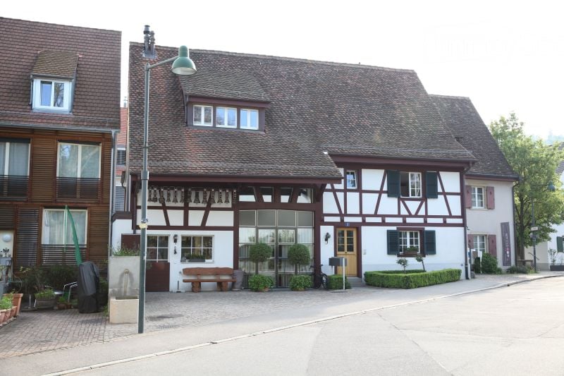 Traditional half-timbered building with a steeply pitched roof, white walls, and dark wooden beams. Features include a central entrance, large windows, and a small bench near the entrance.