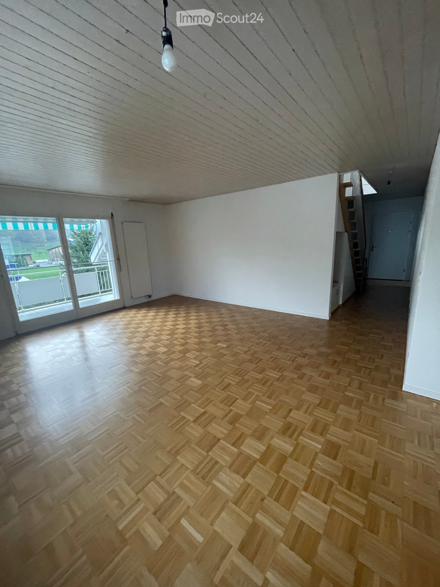 Empty room with wooden herringbone flooring, white walls, and a large window with a view of the balcony and outdoor area.