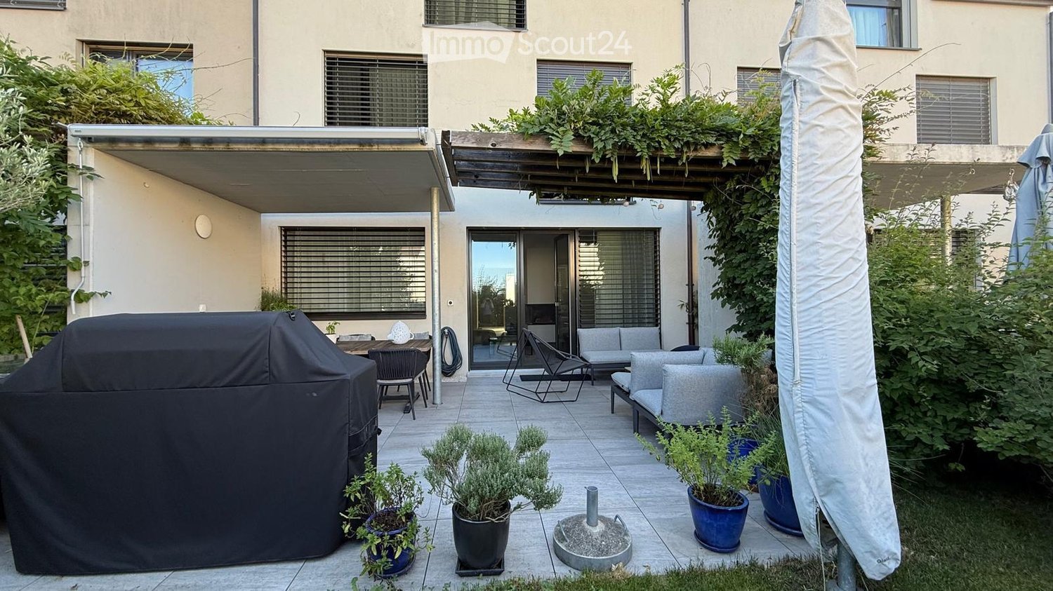 A modern outdoor patio with a covered seating area, featuring a sofa, table, chairs, potted plants, and a large umbrella. The patio is connected to a building with large windows and a covered walkway.