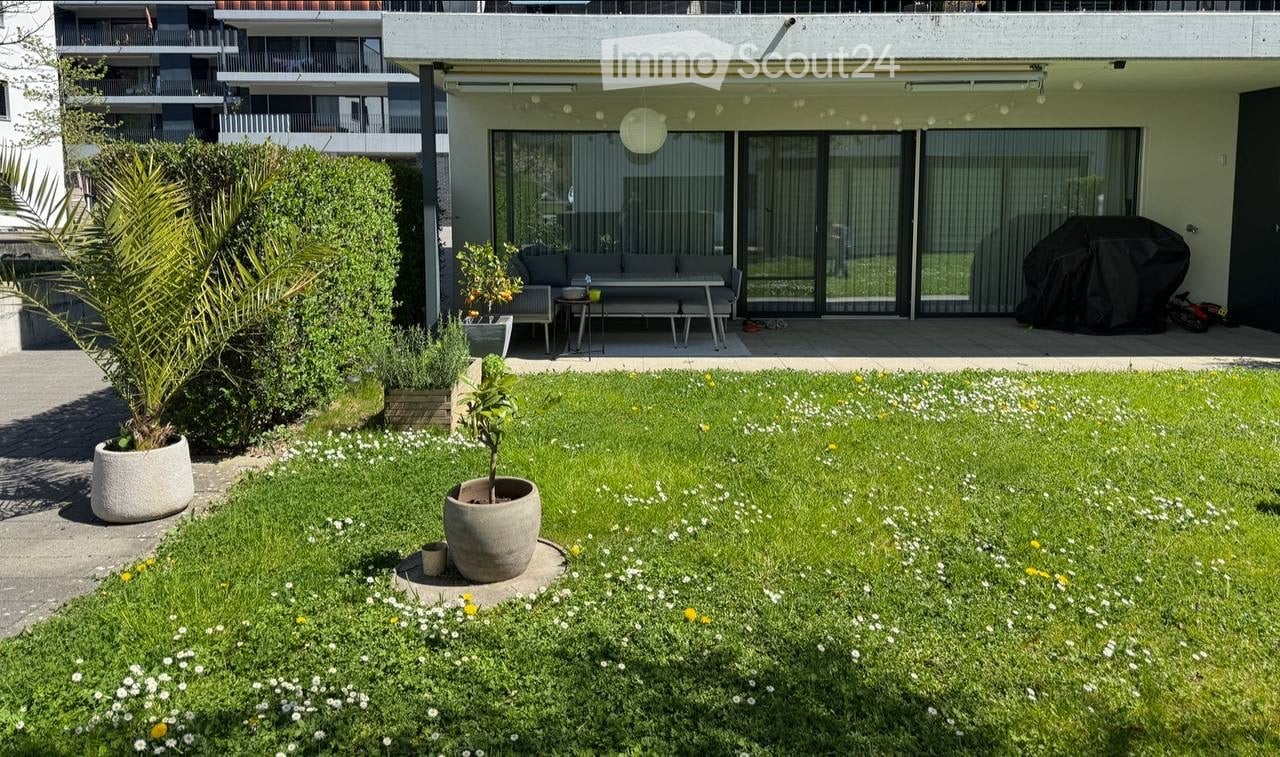 Well-maintained garden with green grass, white flowers, potted plants, and a small seating area with a sofa and table near a modern building.