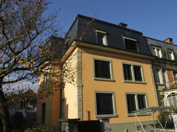 A multi-story residential house with a yellow facade, multiple windows, and a dark roof.
