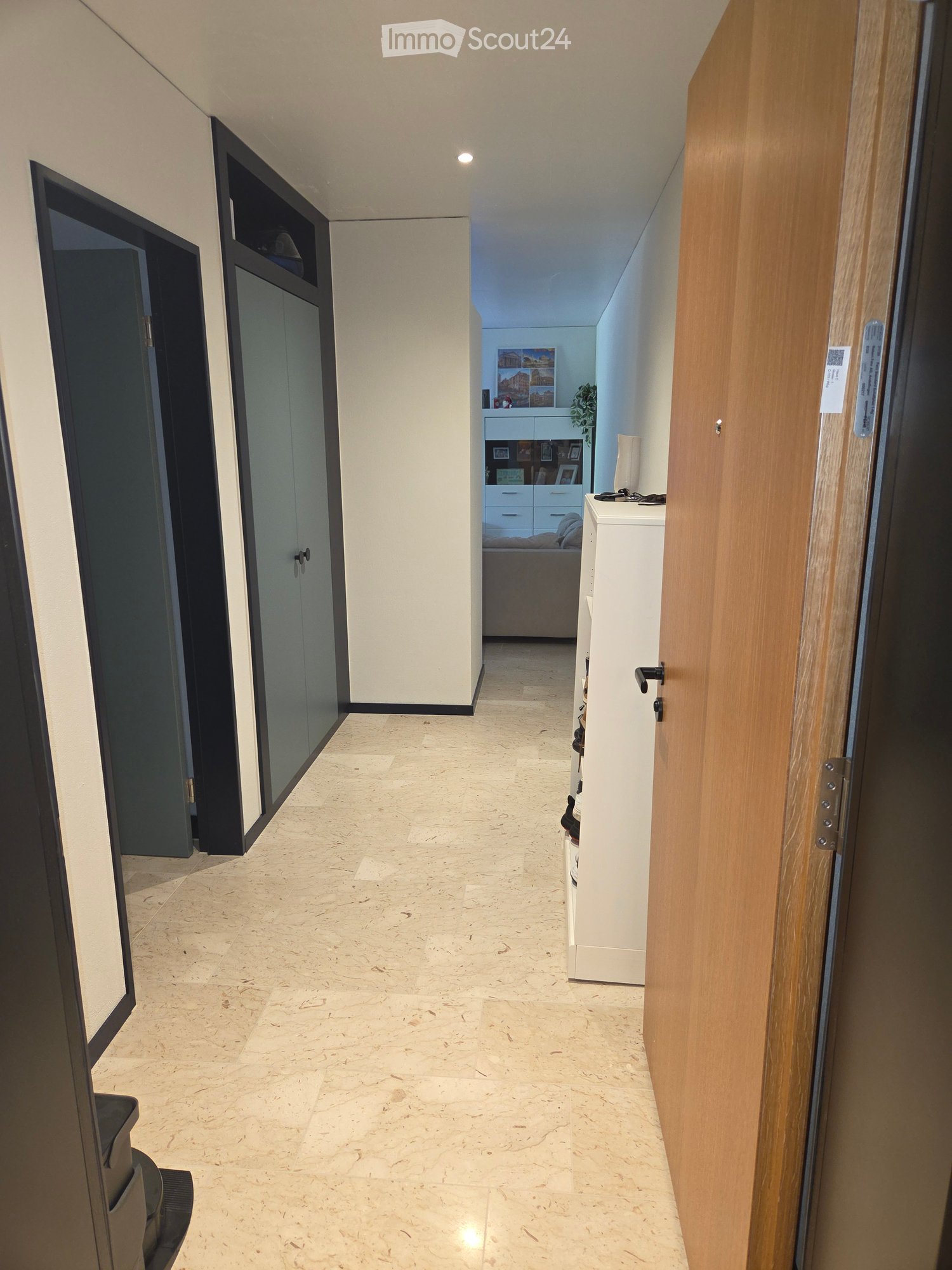Modern hallway with marble flooring, white walls, and several doors leading to different rooms. A small table with a lamp and a plant is visible in the background.