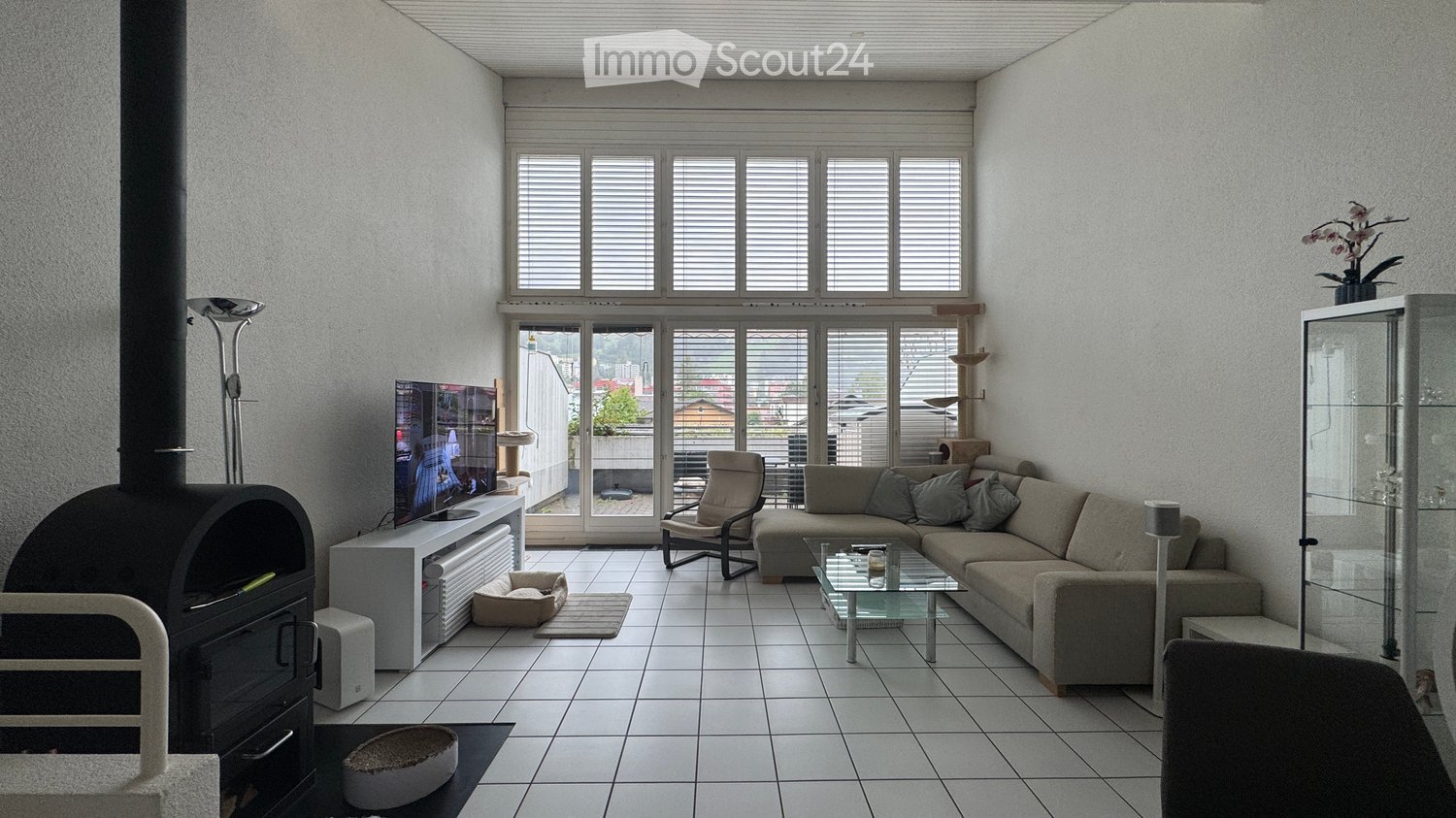 Modern living room with large windows, white tiled floor, grey sectional sofa, glass coffee table, armchair, TV, fireplace, and cat accessories.